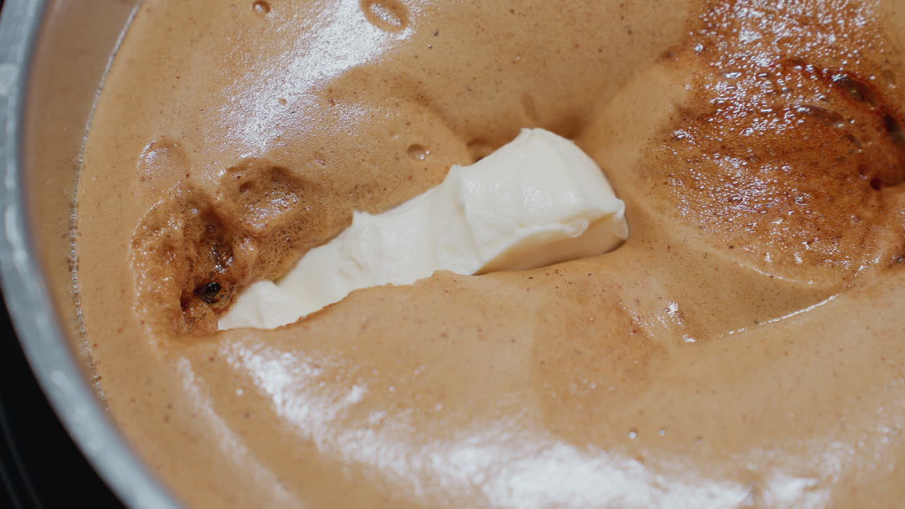 primer plano de mantequilla blanca cayendo en pap marrón caliente, derritiéndose lentamente en la textura espesa y cremosa, la superficie parece aireada, con pequeñas burbujas y ricas variaciones de color