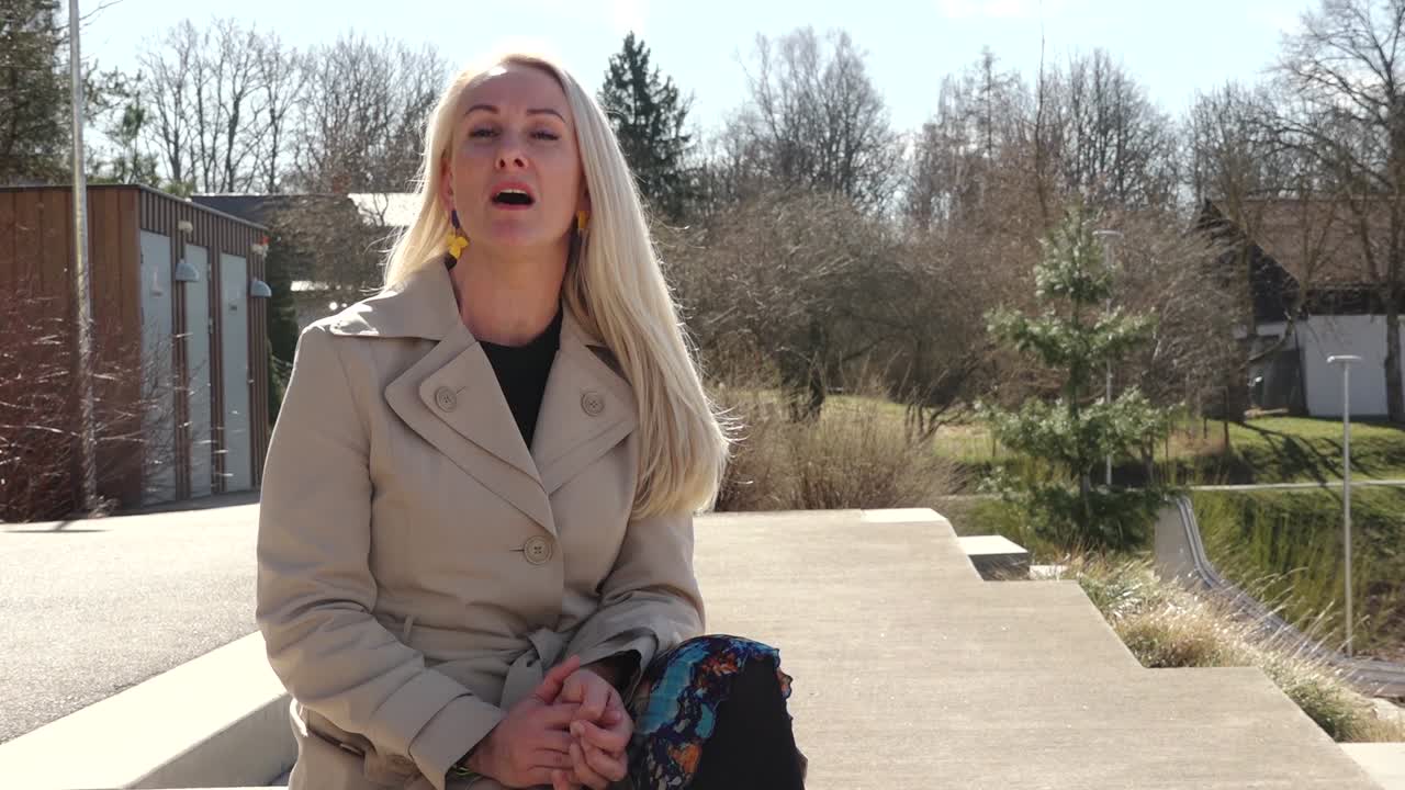 A woman with long blonde hair and colorful earrings smiles confidently while standing outdoors in a beige coat, with trees and buildings in the background.