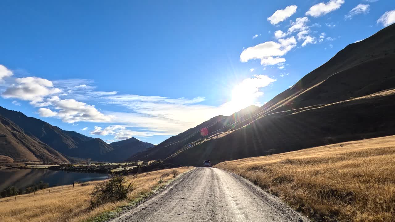 Vehicle travels gravel road in sunlit mountain valley, wide angle, natural light, scenic landscape