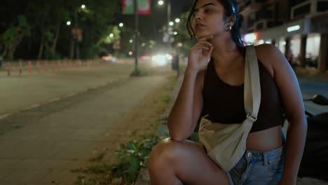 Mujer-De-Moda-En-Cuclillas-En-Una-Acera-Por-La-Noche-Con-Las-Luces-De-La-Ciudad-Detrás