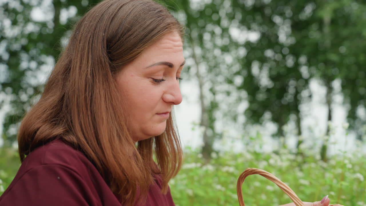 흰색 셔츠를 입은 여성이 자작나무 근처에서 빵 굽는 냄새를 맡고 있어요. 엮은 바구니에서 갓 구운 빵 냄새를 맡으며 생각에 잠긴 표정을 짓고 있고, 배경에는 야생화 초원이 펼쳐져 있으며 평화로운 시골 풍경을 담은 정적이고 친밀한 사진이에요.