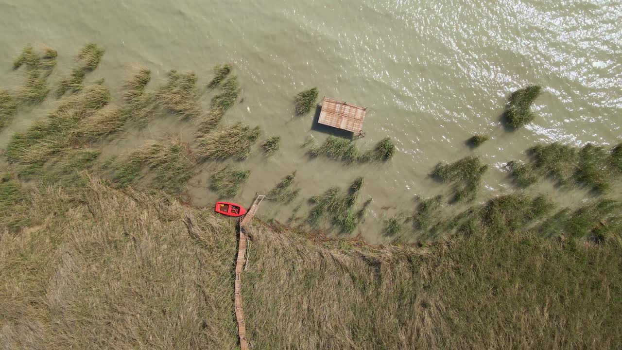 An aerial shot captures a wooden pier extending into a calm lake, with a red boat docked at the end. Tall lake reeds surround the scene.
