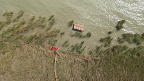 an aerial shot captures a wooden pier extending into a calm lake, with a red boat docked at the end. tall lake reeds surround the scene.
