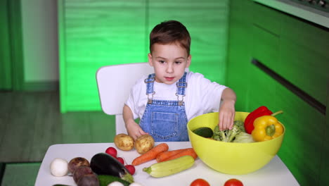 boy exploring veggies in kitchen. a small child sits at a table surrounded by fresh vegetables, curiously examining a large bowl of produce