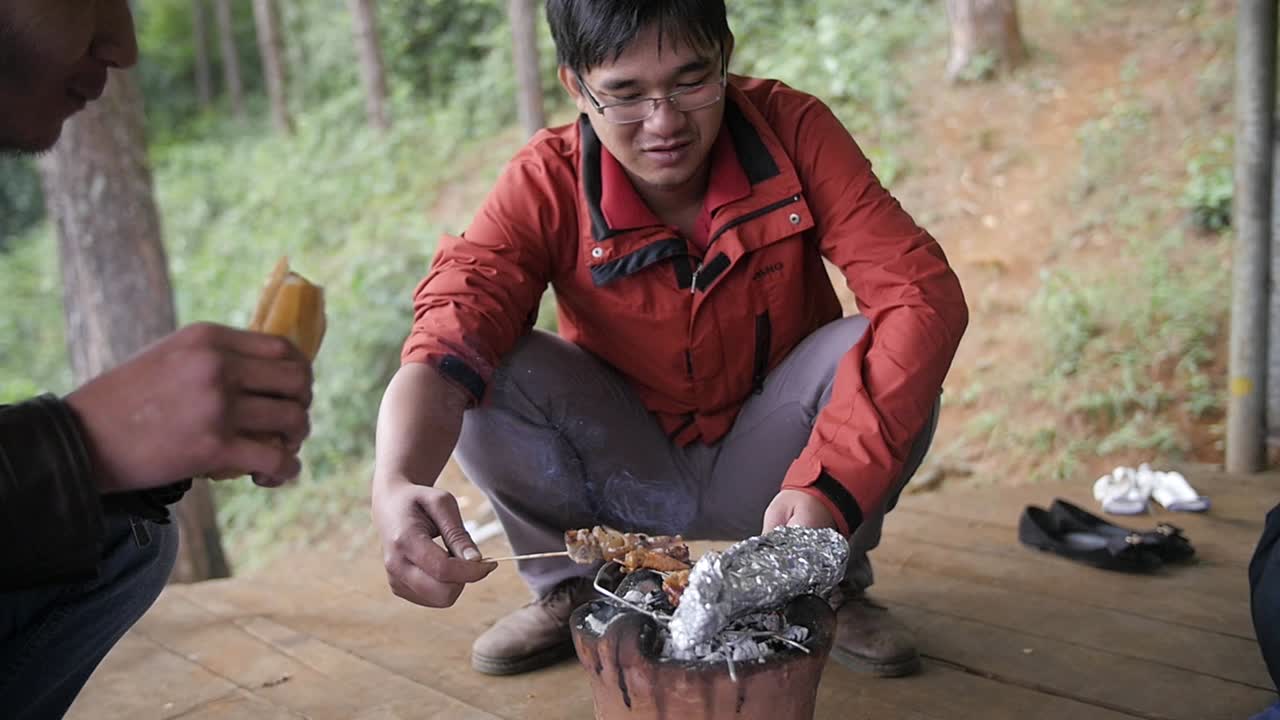Slow motion Vietnamese male crouched with barbecue smoking, Cooking meat on skewer.