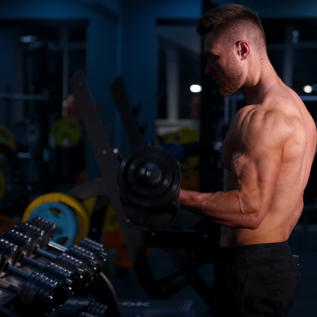 Giovane uomo in forma che solleva manubri e si allena in palestra. Concetto di sport, fitness, sollevamento pesi, bodybuilding, allenamento, atleta, esercizi. Vista laterale.