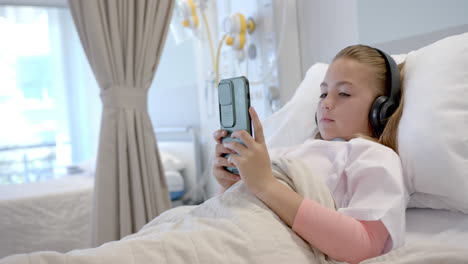 Paciente-Caucásica-En-Cama-De-Hospital-Con-Auriculares-Usando-Smartphone,-Espacio-Para-Copiar,-Cámara-Lenta