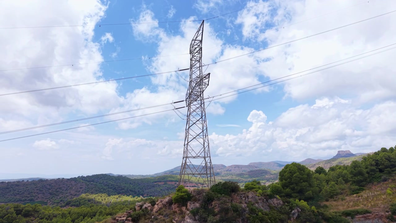 Tall steel electricity pylon standing on a rocky hilltop surrounded by green Mediterranean landscape with mountains and blue cloudy sky in background