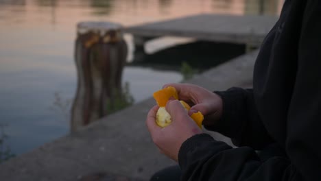 primer plano de un hombre sentado cerca de un muelle durante la puesta de sol y pelando una deliciosa naranja