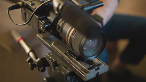 close-up shot of camera tripod mounting plate, cameraman adjusting camera in background, green indoor environment with blurred details, focus on tripod component