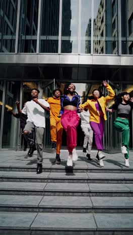 six young dancers in colorful outfits are joyfully jumping down the stairs of a modern glass building, showcasing their energy and enthusiasm