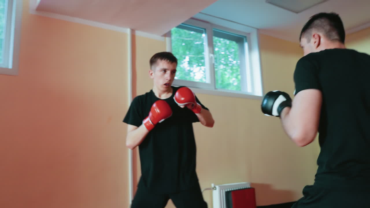 Fighters sparring inside gym, young man in red gloves delivering punch toward opponent in black gloves, practicing attack and defense, showing intensity, focus, energy, agility, and martial arts