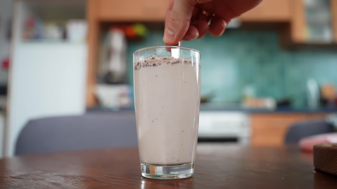 Una mano remueve un vaso alto de leche con chocolate sobre una mesa de madera, con un acogedor fondo de cocina y poca profundidad de campo. Preparación casera de una bebida, primer plano de estilo de vida.