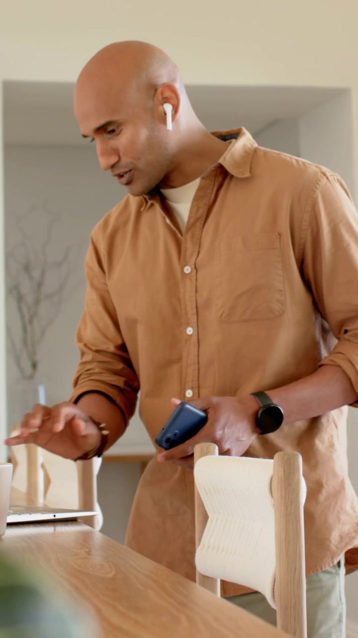 Vertical video: Taking call tan-shirted man at home table, with phone, using laptop with earbuds