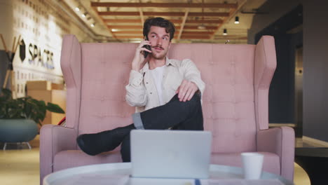 Millennial-white-male-creative-sitting-on-sofa-in-an-office-lobby-making-a-phone-call,-close-up