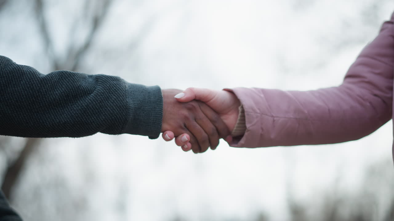 Two people extending hands towards each other for handshake outdoors, one in dark textured hoodie and other in pink winter jacket, symbolizing upcoming greeting, cooperation, agreement, in park setting