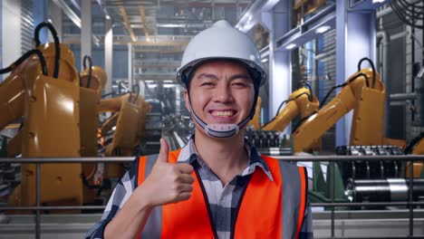 close up of asian male engineer with safety helmet smiling and showing thumbs up gesture to the camera while standing with industrial robotic arms working