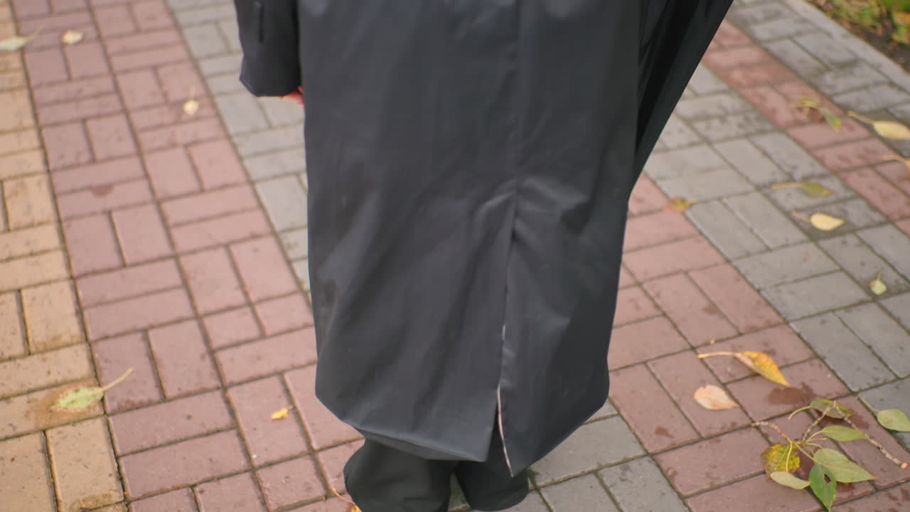 Rear view of person in black coat holding closed black umbrella with wooden handle while walking on wet paved sidewalk outdoors, symbolizing preparation, anticipation, weather protection