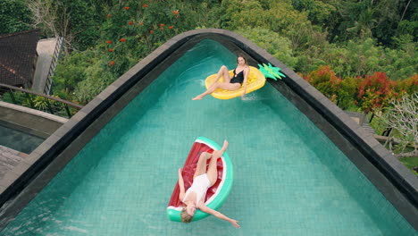 two-women-floating-in-swimming-pool-lying-on-inflatables-best-friends-having-fun-summer-day-on-vacation-at-tropical-hotel-resort-wearing-bikinis-top-view