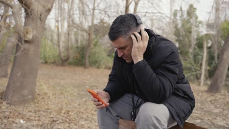 a man sits in the woods scrolling through his mobile phone while listening to music through his headphones