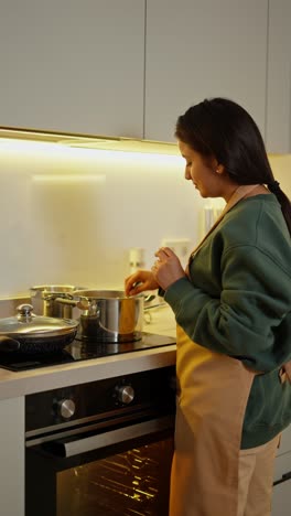 From-behind,-a-happy-brunette-girl-with-Brown-skin-in-a-green-sweater-and-a-beige-apron-prepares-food-using-a-saucepan-while-in-a-modern-kitchen-in-an-apartment-in-the-evening