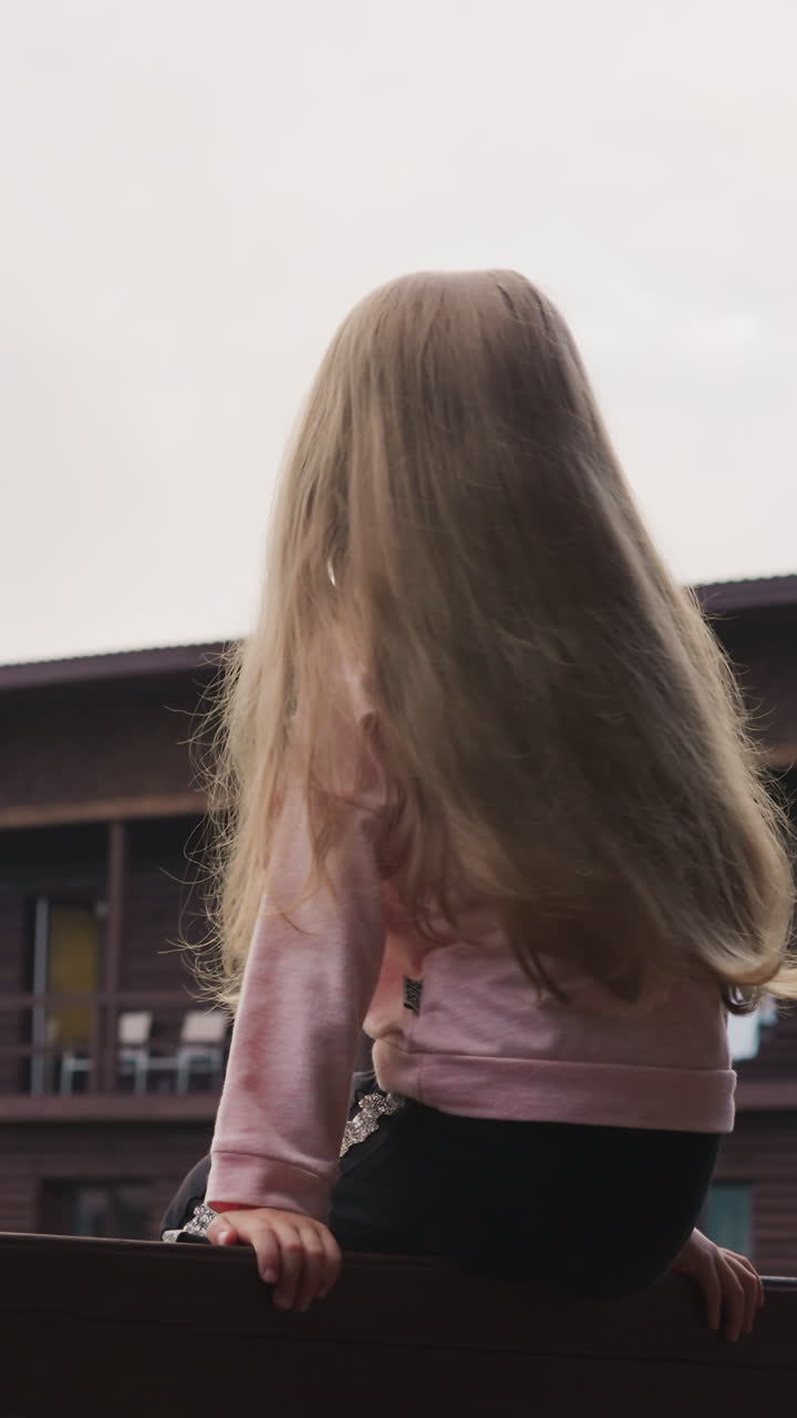 긴 느슨한 머리카락을 가진 귀여운 작은 소녀는 큰 안개 산에 대항하여 에코 호텔 테라스의 나무 레일링에 앉아 있습니다.