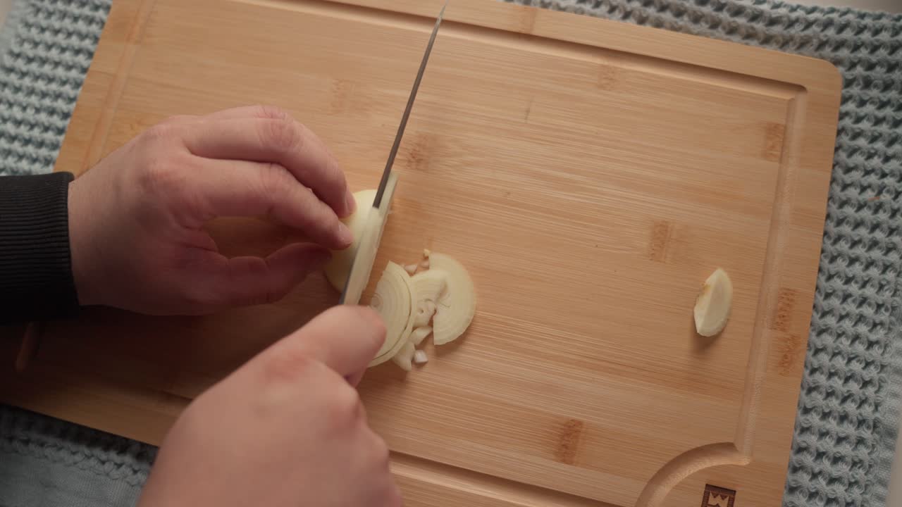Una persona affetta uno spicchio d'aglio con un coltello da cucina su un tagliere di legno durante la preparazione del pasto. Il primo piano evidenzia il processo di cottura e la preparazione degli ingredienti in una cucina domestica.