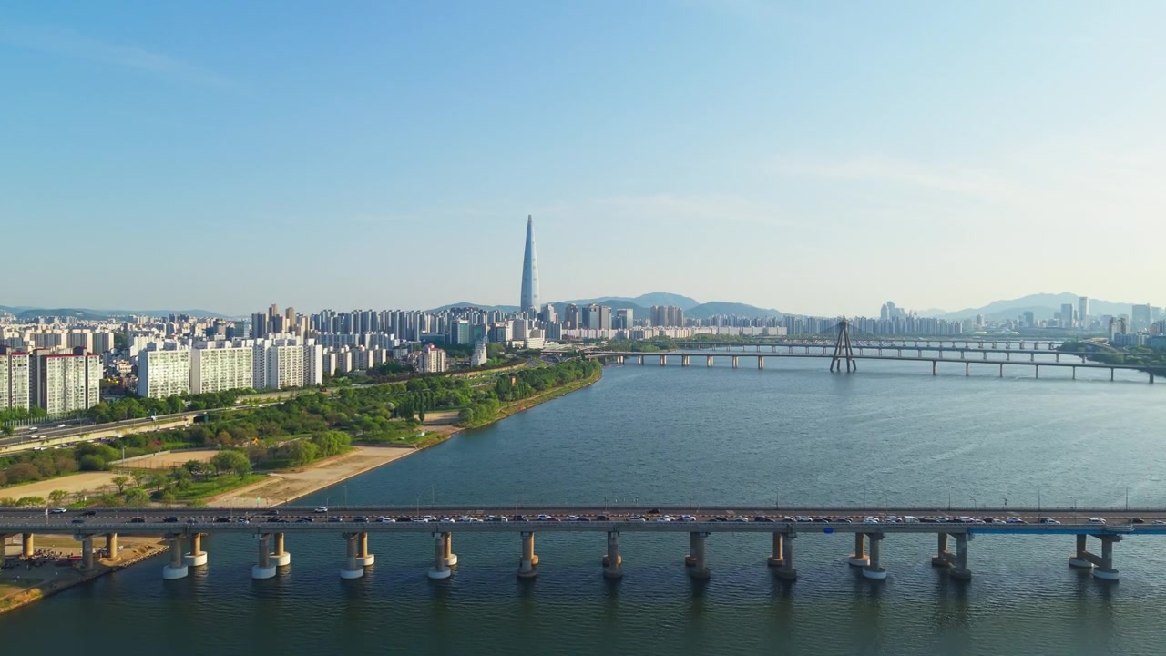 Sorvolo aereo lungo il fiume Han a Seul che passa i ponti Gwangjin e Cheonho, con l'iconica Lotte World Tower che si erge sopra lo skyline della città sotto un cielo diurno sereno.