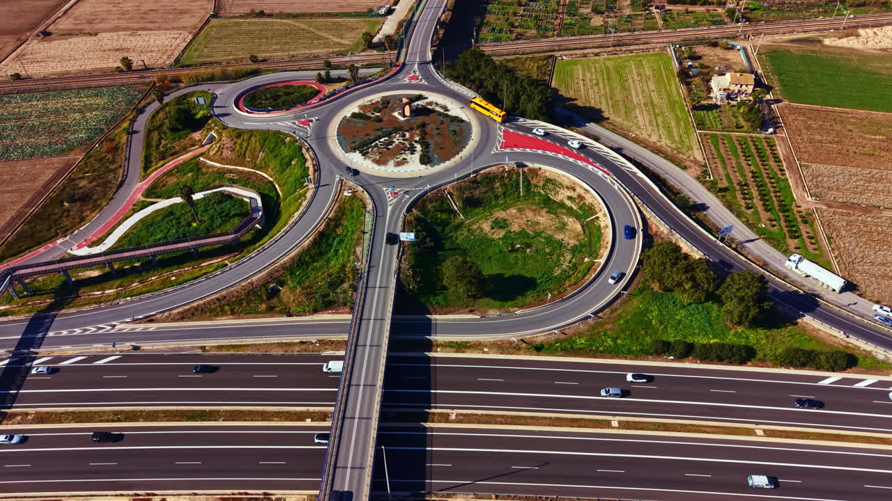 Roadway system shows circular interchange with fields and buildings nearby in daytime