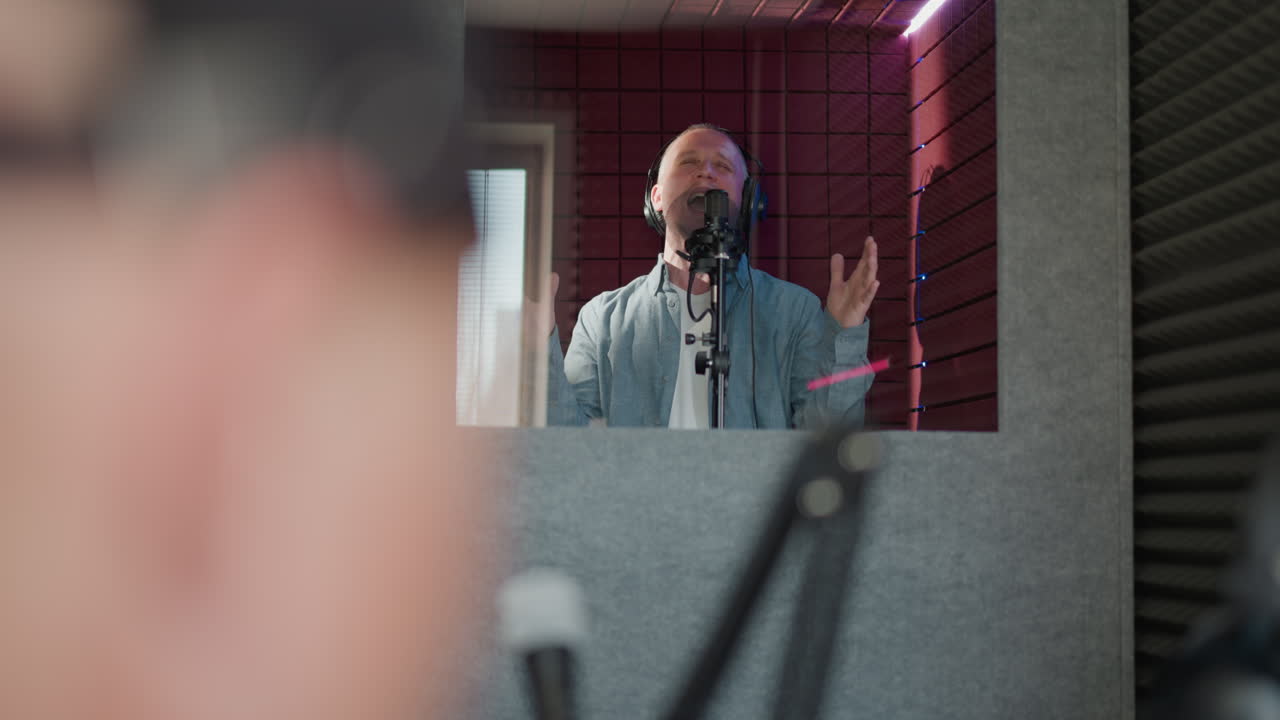 el hombre canta apasionadamente en un estudio de grabación, con auriculares negros y una camisa azul de manga larga. la escena se filma a través de un espejo, un gesto de la mano, en un fondo rojo con paneles acústicos