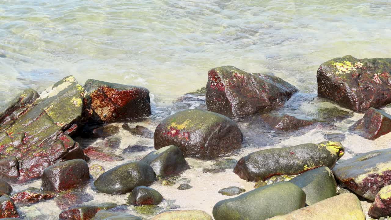 Waves gently crash over red algae-covered rocks on a sunny Phuket shoreline, creating a dynamic and serene coastal scene