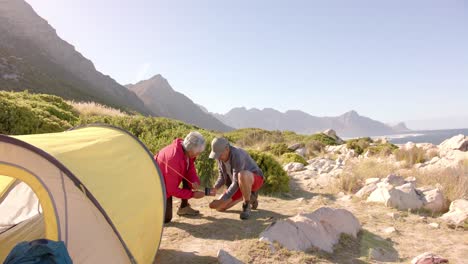 Happy-senior-biracial-couple-preparing-tent-in-mountains-on-sunny-day,-in-slow-motion