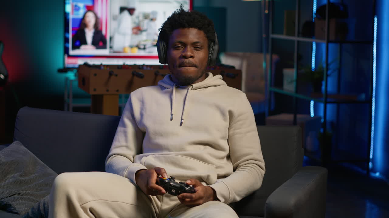 African american man playing shooter videogame in his loft apartment, holding controller for leisure activity. Male gamer participating in internet multiplayer game at night. Camera A.