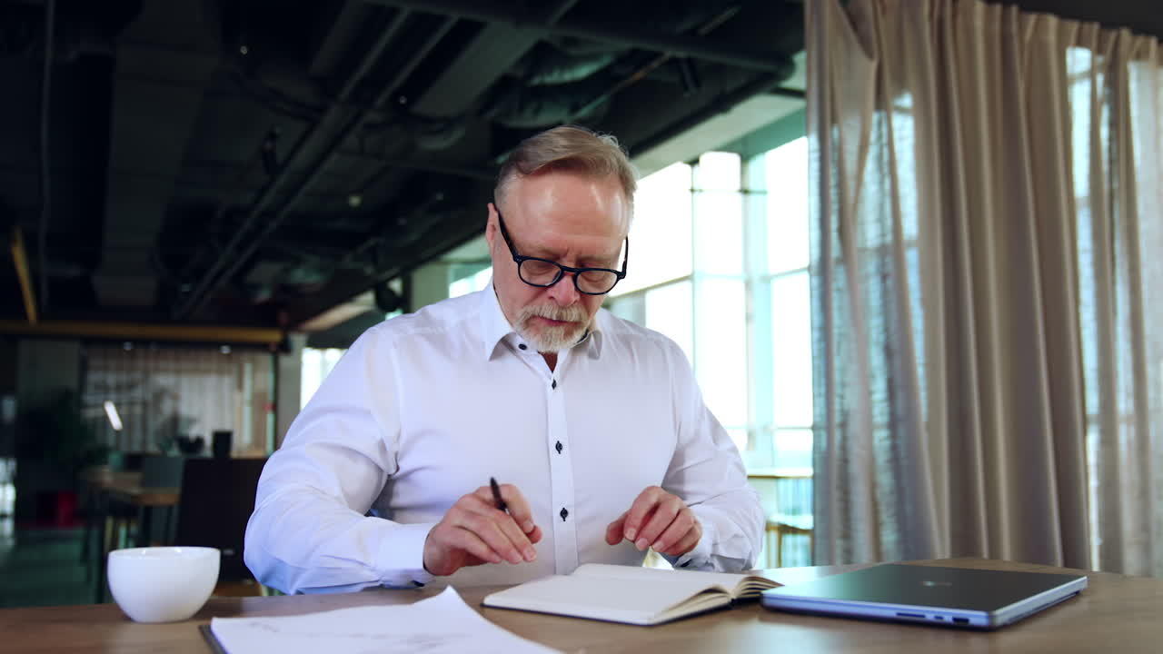 Serious man in white shirt and glasses drinks tea of coffee from a cup. Businessman turns the pages of the paper notebook and look aside.