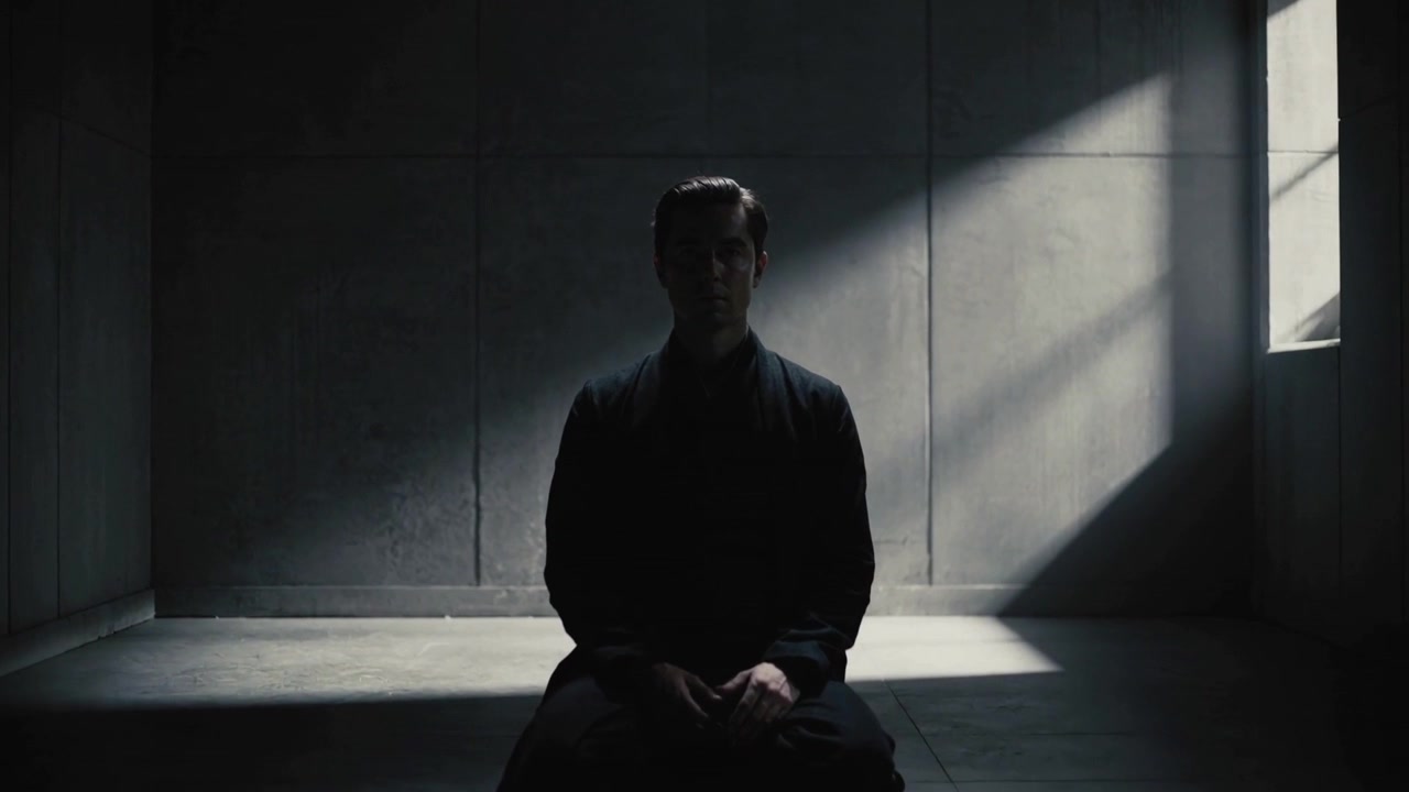 Camera zooming revealing kneeling man in concrete room under high window, dark robe revealing face