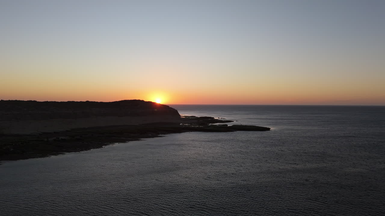 Stunning 4K 60fps aerial shot of Puerto Madryn’s ocean at sunrise, with vibrant sky colors, shadowed cliffs, and peninsula. Distant horizon on scene.