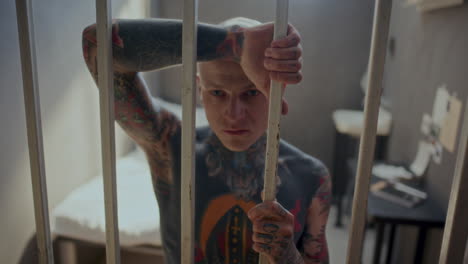 bald tattooed inmate holding prison cell bars and looking away with anger