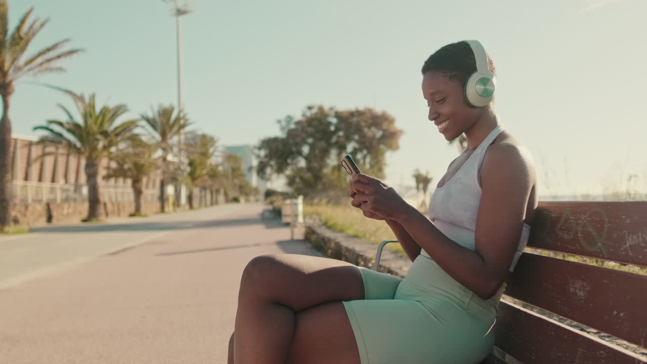 Giovane donna afroamericana con le cuffie che usa lo smartphone su una panchina del parco