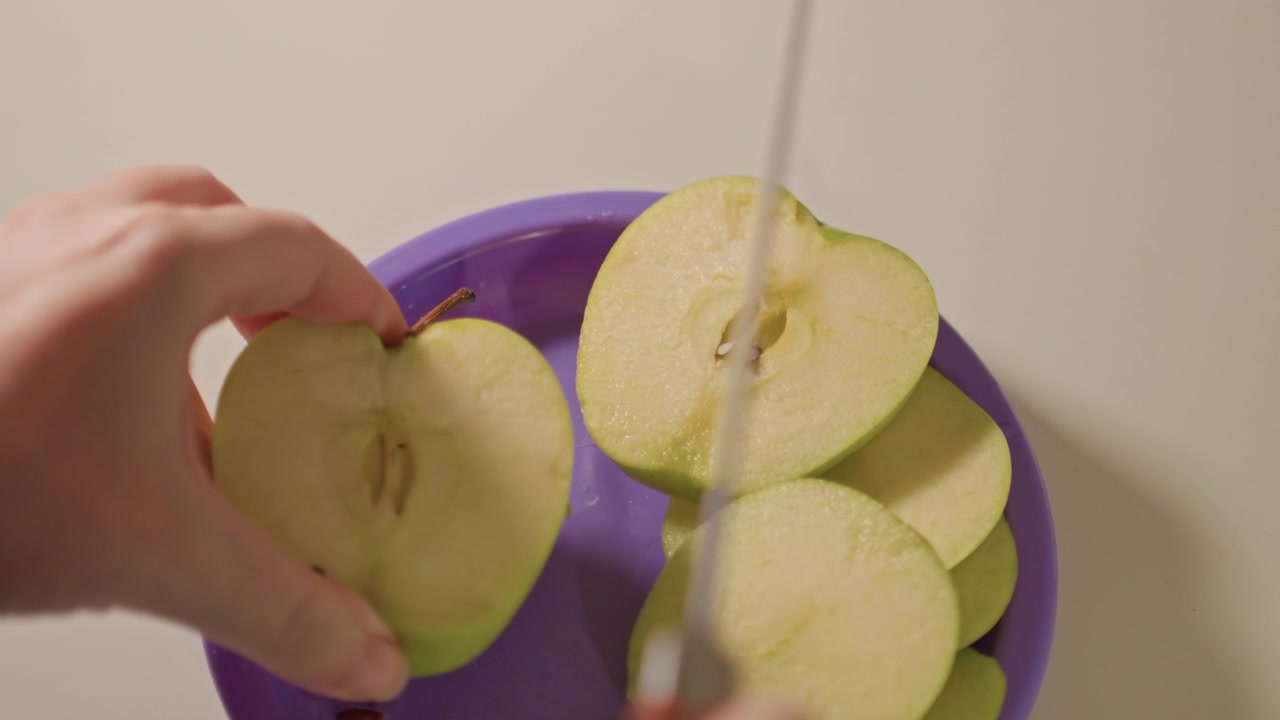 Primo piano di mani che affettano mele verdi a pezzi sottili. Un coltello con dettagli neri e rossi taglia il frutto, rivelando i semi. Lo sfondo semplice enfatizza i dettagli della preparazione del cibo