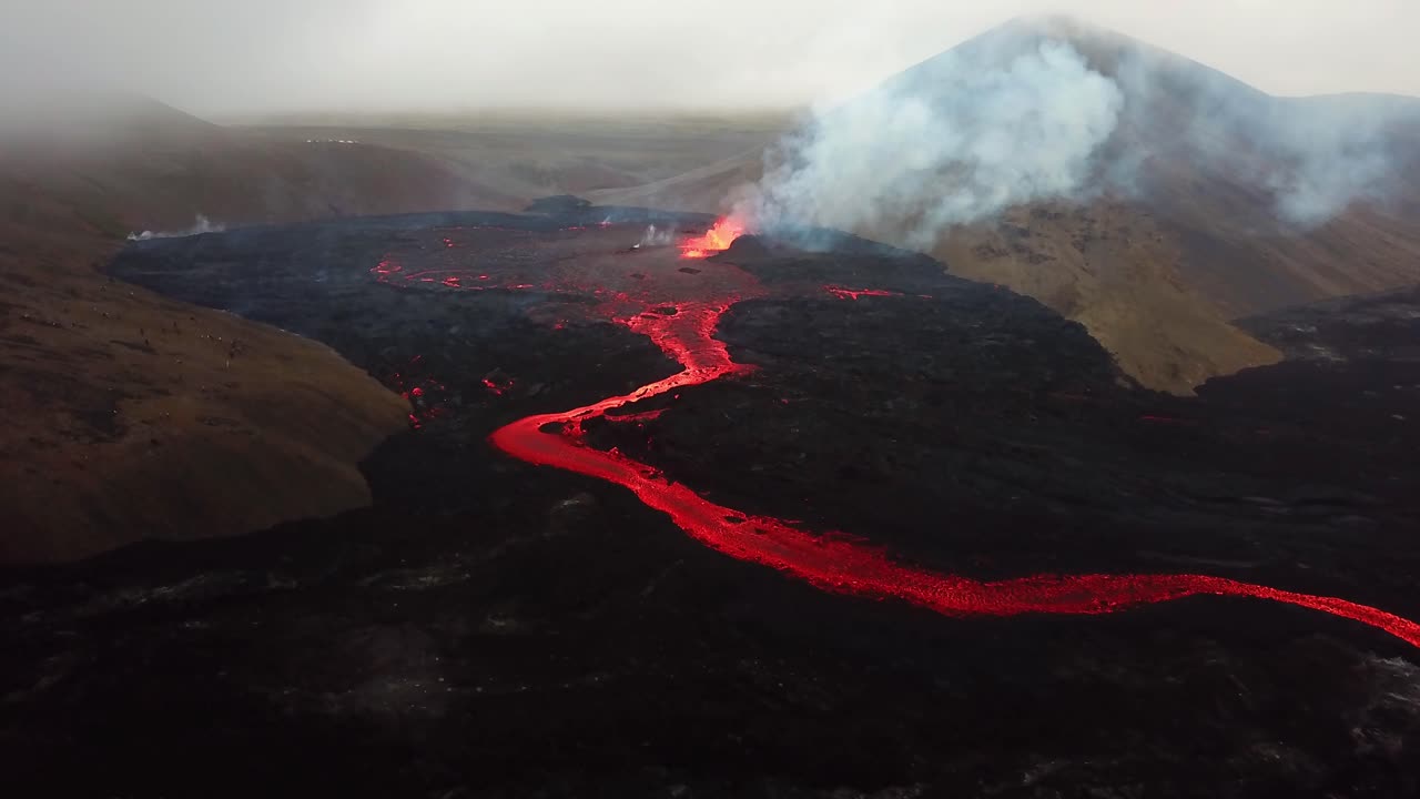 Aerial landscape view of Fagradalsfjall volcano erupting with lava flowing across the Meradalir valley floor, and smoke coming out
