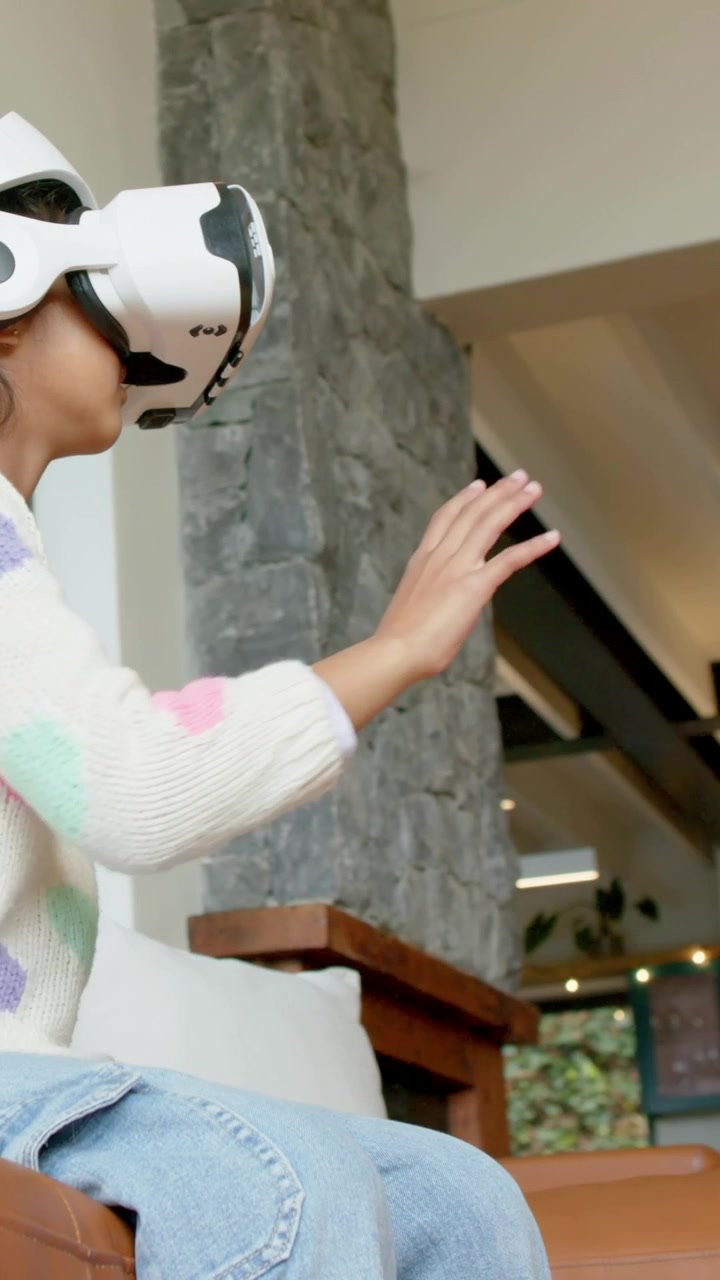 Video verticale: Ragazza con visore VR si allunga seguendo un oggetto che appare sul divano, maglione, spazio per il testo.
