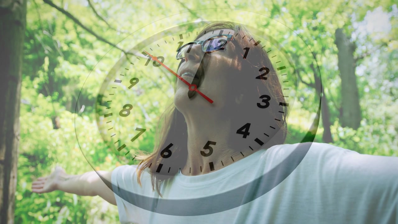 Mujer madura de pie con los brazos abiertos y la cabeza hacia atrás en el bosque, un reloj girando sobre ella ilustrando el bienestar.