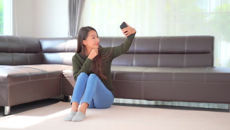 Beautiful-and-cute-Asian-young-woman-smiling-cheerfully-and-posing-to-the-smartphone-camera-while-taking-selfie-photos-or-video-on-front-camera-in-home-interior