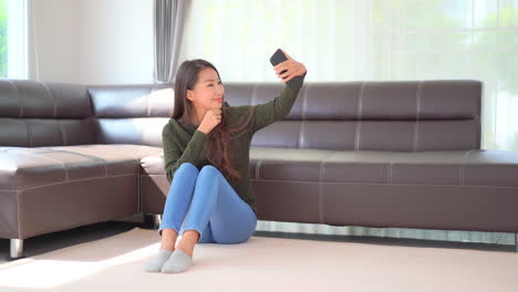 아름답고 귀여운 아시아 젊은 여성은 유쾌하게 웃고 스마트폰 카메라에 포즈를 취하면서 셀카 사진이나 비디오를 집 내부의 전면 카메라로 찍습니다.