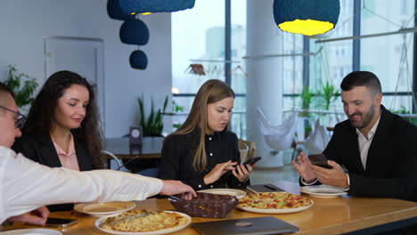 business people having lunch and discussing work in smartphones. colleagues talking about project and eating pizza together in spacious office during lunch break.