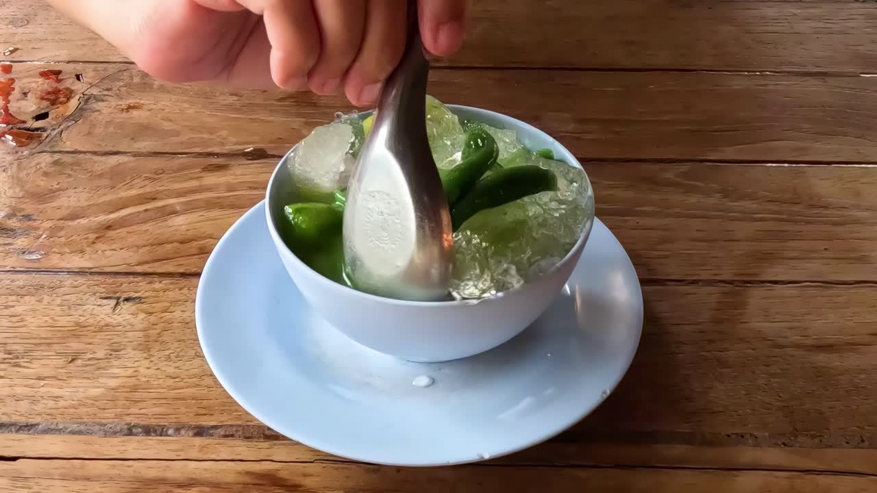 Close-up of hands mixing green shaved ice with various toppings in a bowl on a wooden table.
