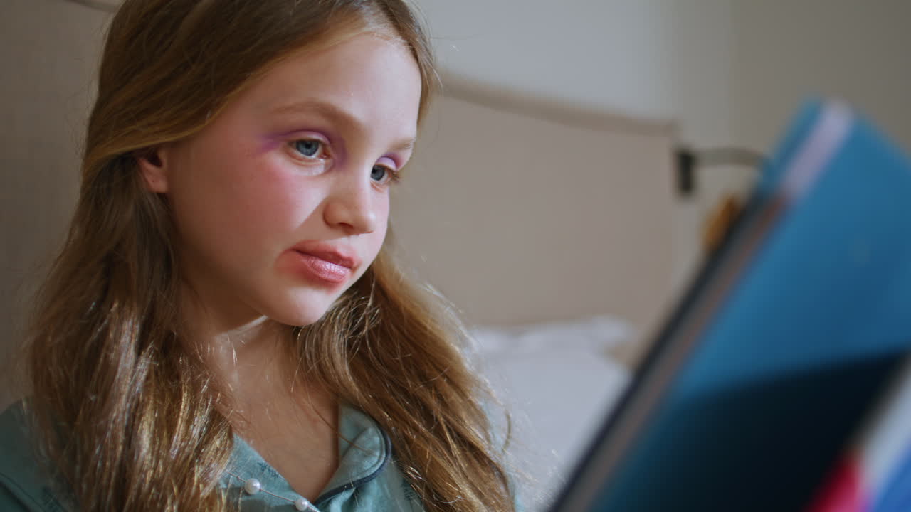 Baby girl flipping book pages sitting bed at home closeup. Child with makeup
