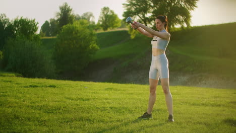 una mujer deportiva en un parque en cuclillas con pesas en las manos al atardecer. entrenamiento con pesas al aire libre. fitness en un parque público en verano para los muslos y las piernas.