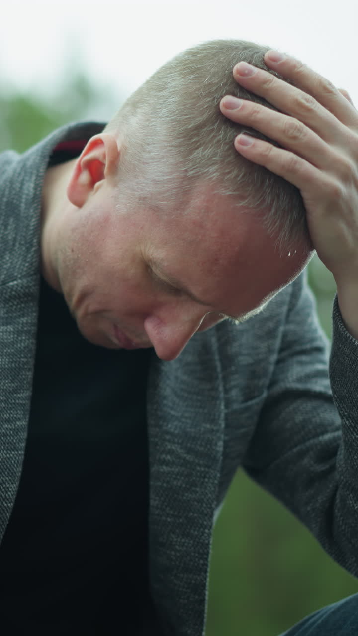 una vista hacia abajo de un hombre con una chaqueta gris y pantalones vaqueros azules, usando zapatos de lona, manejando una pistola mientras está sentado al lado de una vía ferroviaria, su cabeza está inclinada, y una mano está en su cabeza