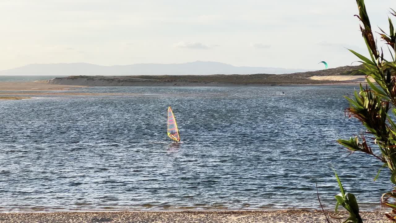 toma cinematográfica en cámara lenta de un windsurfista en primer plano en lagoa de albufeira, portugal, europa y un kitesurfista en el fondo en un día soleado con mucho viento